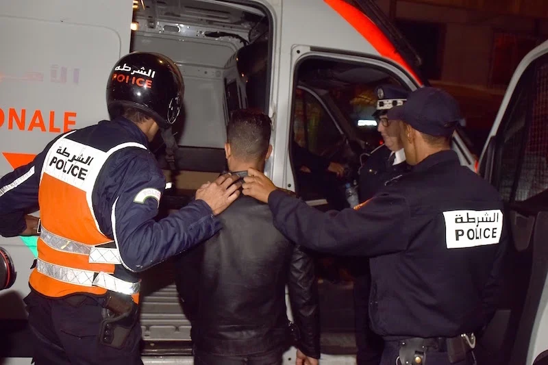 Police officers escort a restrained man toward a police van at night, placing hands on his shoulders as he enters the vehicle (Arabic POLICE text on uniforms).