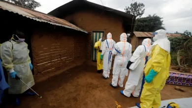 Healthcare workers in full PPE (yellow/white suits, masks) entering a mud-brick house, one spraying disinfectant at the doorway outside.