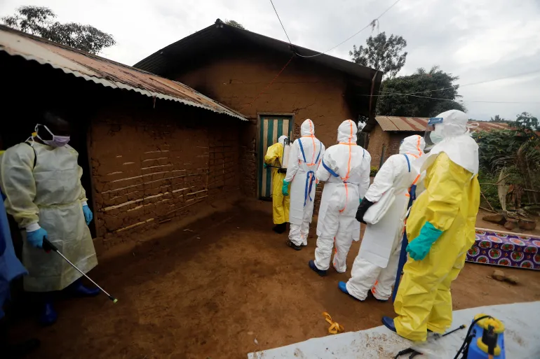 Healthcare workers in full PPE (yellow/white suits, masks) entering a mud-brick house, one spraying disinfectant at the doorway outside.