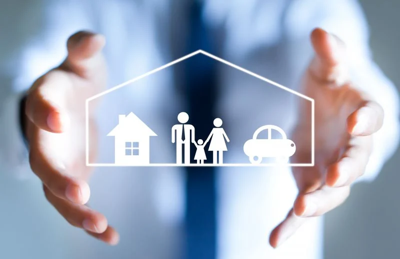 Hands surround a white outline of a house, family, and car inside a protective shape, symbolizing home insurance protection for a family.