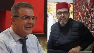 Two men seated side by side: left man smiles in a light blue shirt and tie, right man wears a red fez and black vest against a patterned backdrop.