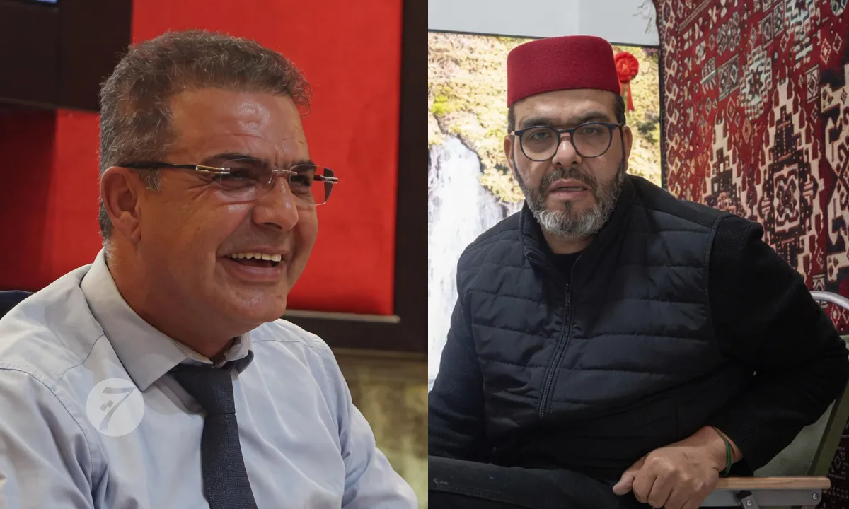 Two men seated side by side: left man smiles in a light blue shirt and tie, right man wears a red fez and black vest against a patterned backdrop.