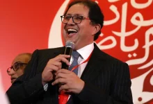 Man in a dark suit and glasses speaks into a microphone at a red-themed event, smiling broadly.