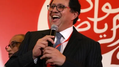 Man in a dark suit and glasses speaks into a microphone at a red-themed event, smiling broadly.