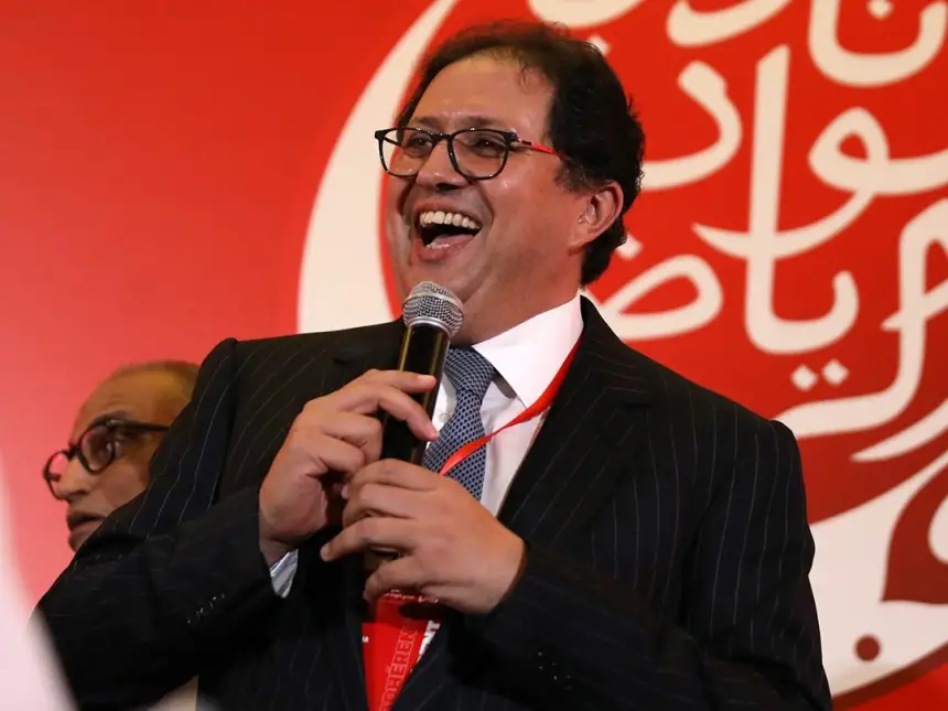Man in a dark suit and glasses speaks into a microphone at a red-themed event, smiling broadly.
