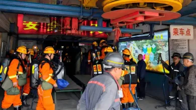 Group of workers in orange uniforms and yellow helmets performing an indoor rescue drill among heavy machinery and blue rails, with Chinese signage nearby.