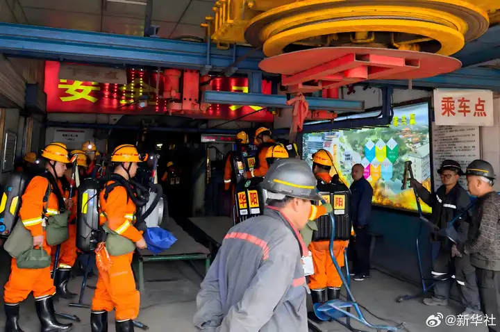 Group of workers in orange uniforms and yellow helmets performing an indoor rescue drill among heavy machinery and blue rails, with Chinese signage nearby.