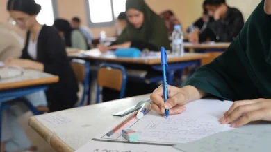 Student in a green hijab writes with a blue pen at a desk in a classroom setting