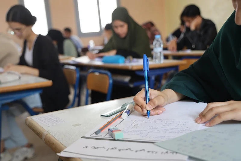 Student in a green hijab writes with a blue pen at a desk in a classroom setting