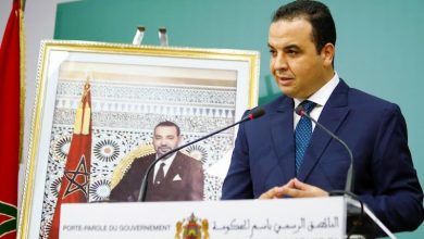 Man in a dark blue suit speaks at a podium with microphones, a framed portrait behind him, and a flag to the side.