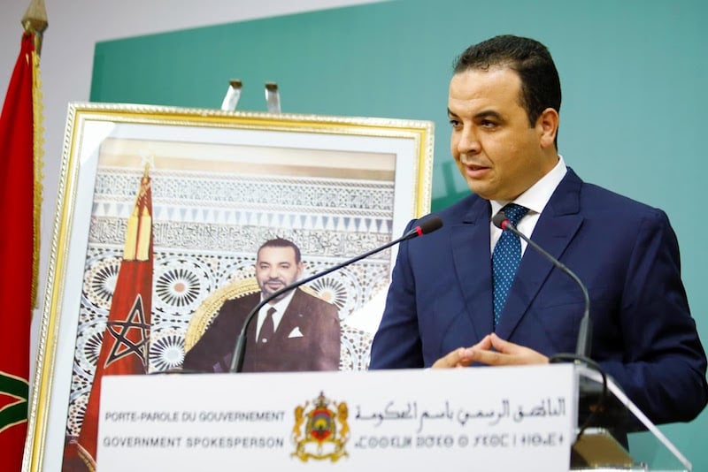 Man in a dark blue suit speaks at a podium with microphones, a framed portrait behind him, and a flag to the side.