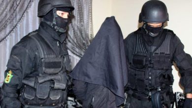 Two masked armed security personnel escort a person in a dark hooded cloak.
