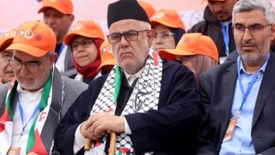 Group of men at a political event wearing orange hats, centered on a man with a black cap, glasses, a patterned scarf, and a cane.