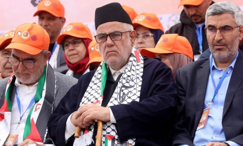 Group of men at a political event wearing orange hats, centered on a man with a black cap, glasses, a patterned scarf, and a cane.