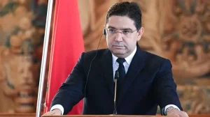Man in a dark suit delivering a speech at a podium with a microphone, in front of a red flag.