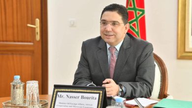 bourita2022 | MCG24 | موقع إلكتروني مغربي شامل متجدد على مدار الساعة Man in a dark suit and pink tie sits at a wooden conference table, with a Moroccan flag in the background and a nameplate visible on the desk.