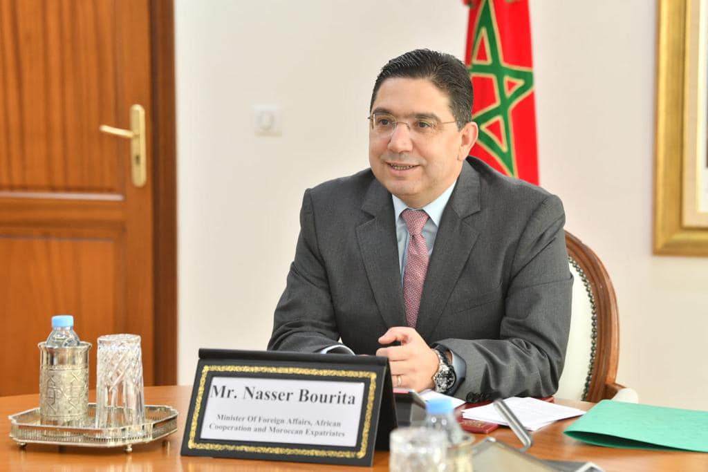 bourita2022 | MCG24 | موقع إلكتروني مغربي شامل متجدد على مدار الساعة Man in a dark suit and pink tie sits at a wooden conference table, with a Moroccan flag in the background and a nameplate visible on the desk.