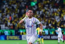 c | MCG24 | موقع إلكتروني مغربي شامل متجدد على مدار الساعة Soccer player in a white jersey with purple stripes celebrates with his hand near his ear on a crowded stadium field.