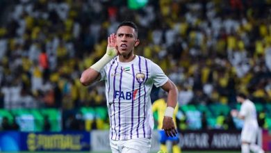 Soccer player in a white jersey with purple stripes celebrates with his hand near his ear on a crowded stadium field.