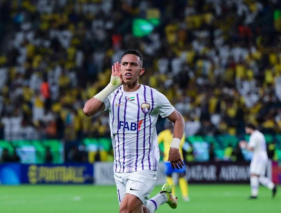 c | MCG24 | موقع إلكتروني مغربي شامل متجدد على مدار الساعة Soccer player in a white jersey with purple stripes celebrates with his hand near his ear on a crowded stadium field.