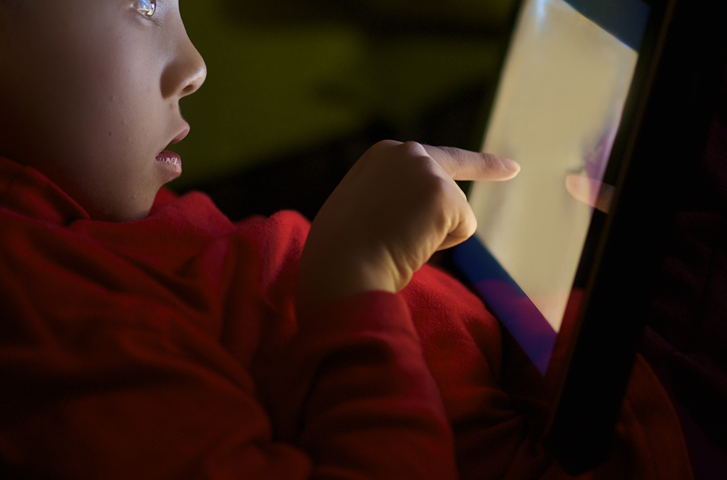 Child in a red shirt touching a glowing tablet screen in low light, profile visible