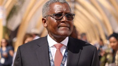 Older man in a dark suit and pink tie, wearing glasses, at an outdoor formal event with a lanyard around his neck.