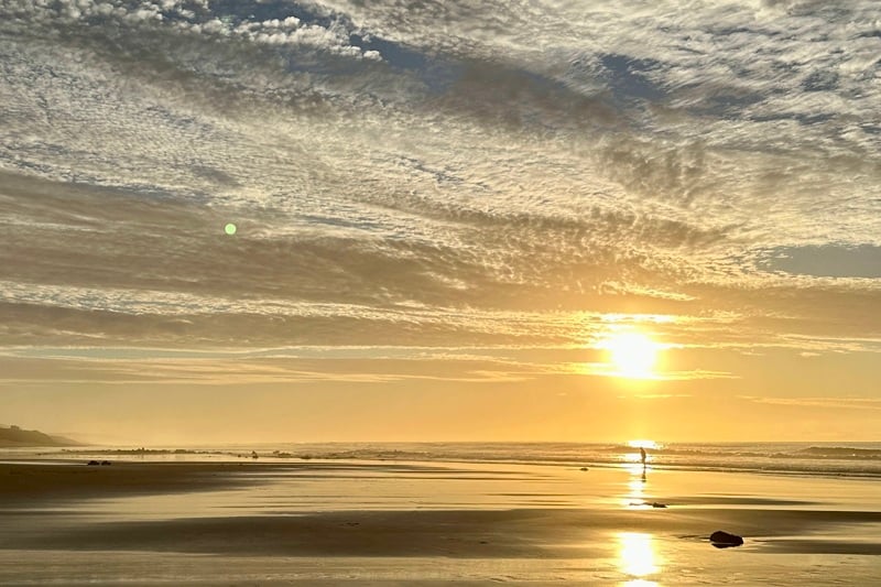 Sunset over a calm beach with reflective wet sand and a lone figure walking along the shoreline.