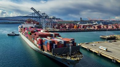 Container ship loaded with colorful shipping containers at a busy port with large gantry cranes in the background.