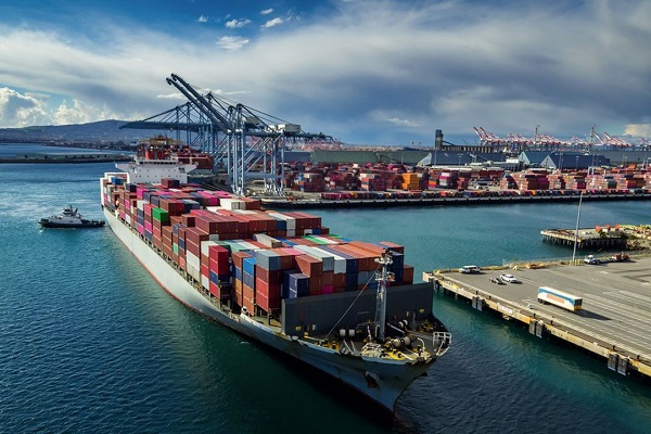 Container ship loaded with colorful shipping containers at a busy port with large gantry cranes in the background.