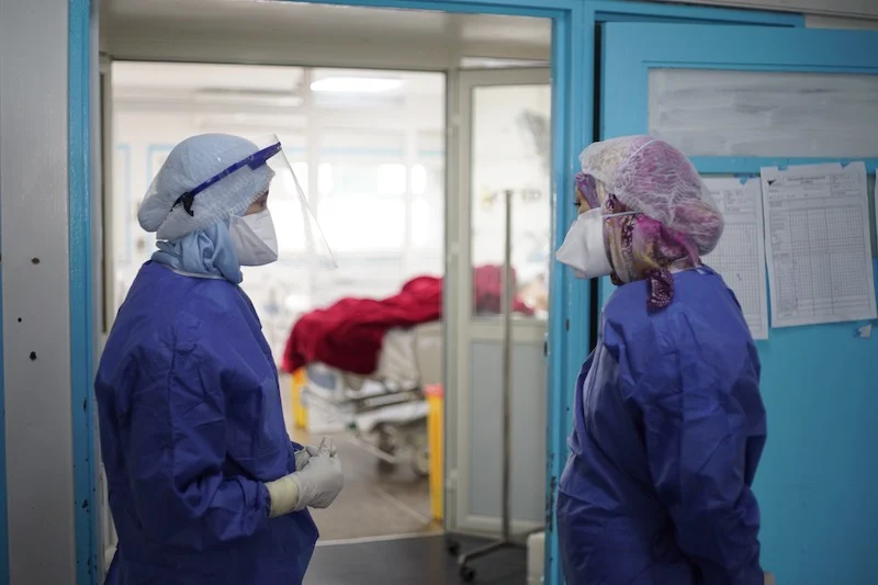 Two healthcare workers in full PPE stand in a hospital doorway, facing each other with a patient bed visible in the background.