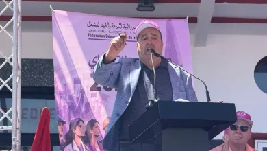 Man speaking at a stage podium with a microphone, wearing a pink cap and gray blazer, purple banner in the background.