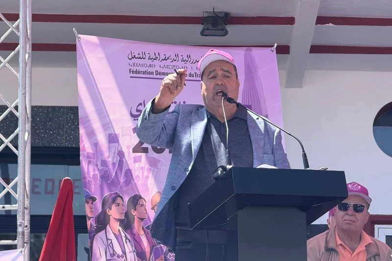 Man speaking at a stage podium with a microphone, wearing a pink cap and gray blazer, purple banner in the background.