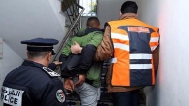 Police officer escorts a handcuffed man up a stairwell, with a person in an orange safety vest nearby.