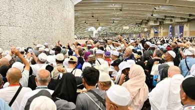 Large crowd of pilgrims gathered along a wall indoors, many wearing white robes and hats, raising hands.