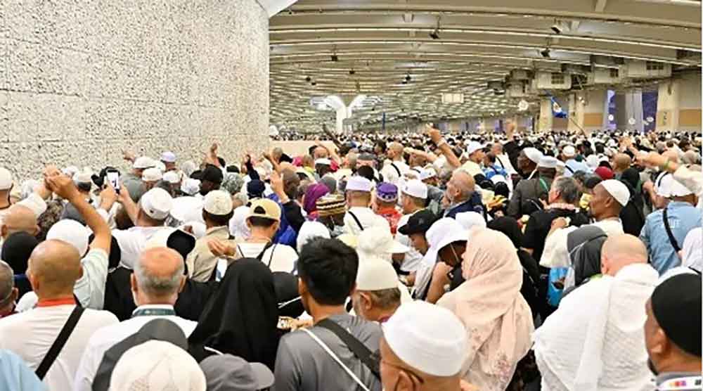 Large crowd of pilgrims gathered along a wall indoors, many wearing white robes and hats, raising hands.