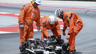 Three orange-clad track marshals inspect a crashed motorcycle on a race track, assessing the damage and ensuring safety.