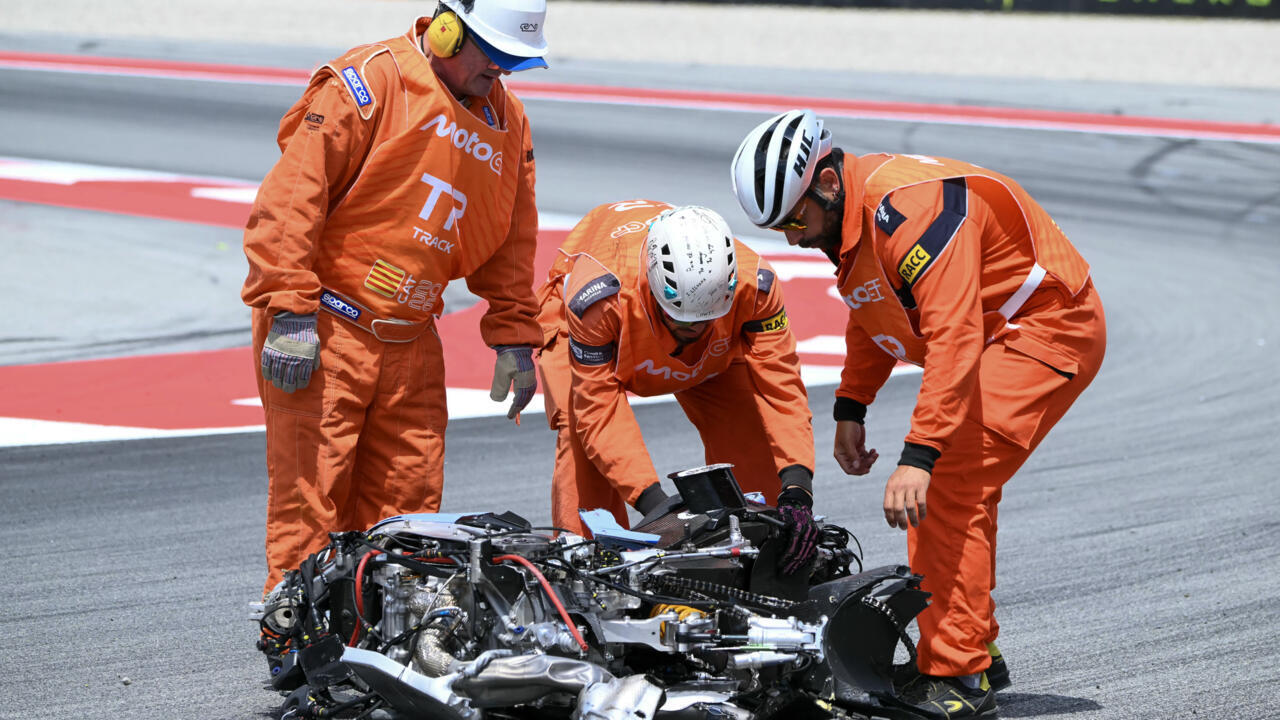 Three orange-clad track marshals inspect a crashed motorcycle on a race track, assessing the damage and ensuring safety.