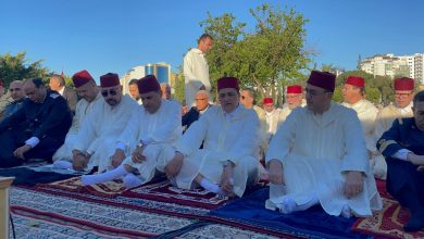 Group of men in white traditional robes and red fez hats sit on prayer carpets outdoors, likely participating in a Muslim prayer gathering in a park with trees and buildings in the background.