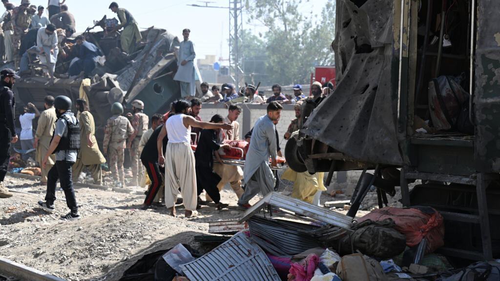 Crowd of people and rescue workers carry a person on a stretcher through wrecked rail cars and debris after a disaster scene.