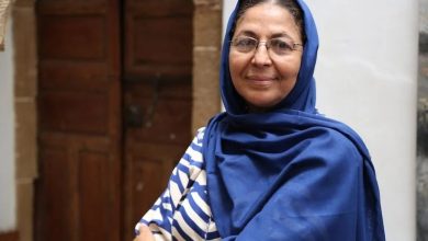 Woman wearing a blue hijab and striped shirt stands outdoors, smiling at the camera.