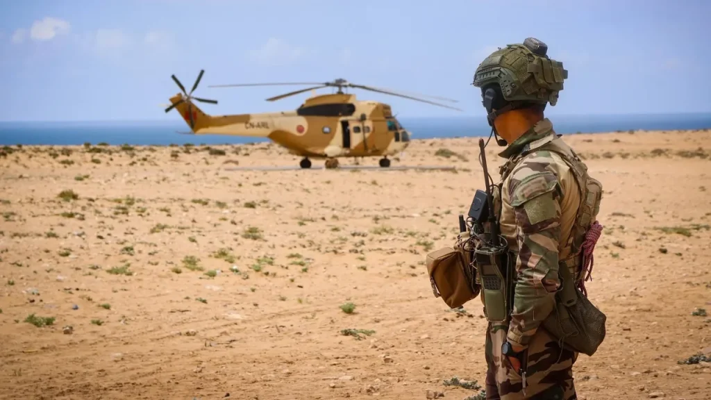 Soldier in full combat gear watches a tan military helicopter on a sandy airstrip with the ocean in the distance.