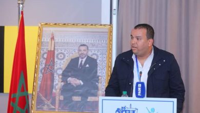 Speaker at a podium with a blue mic, framed portrait behind him, and a Moroccan flag to the left.