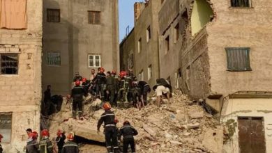 Emergency responders in orange helmets search through a collapsed building's rubble between beige apartment blocks, with onlookers atop the walls.