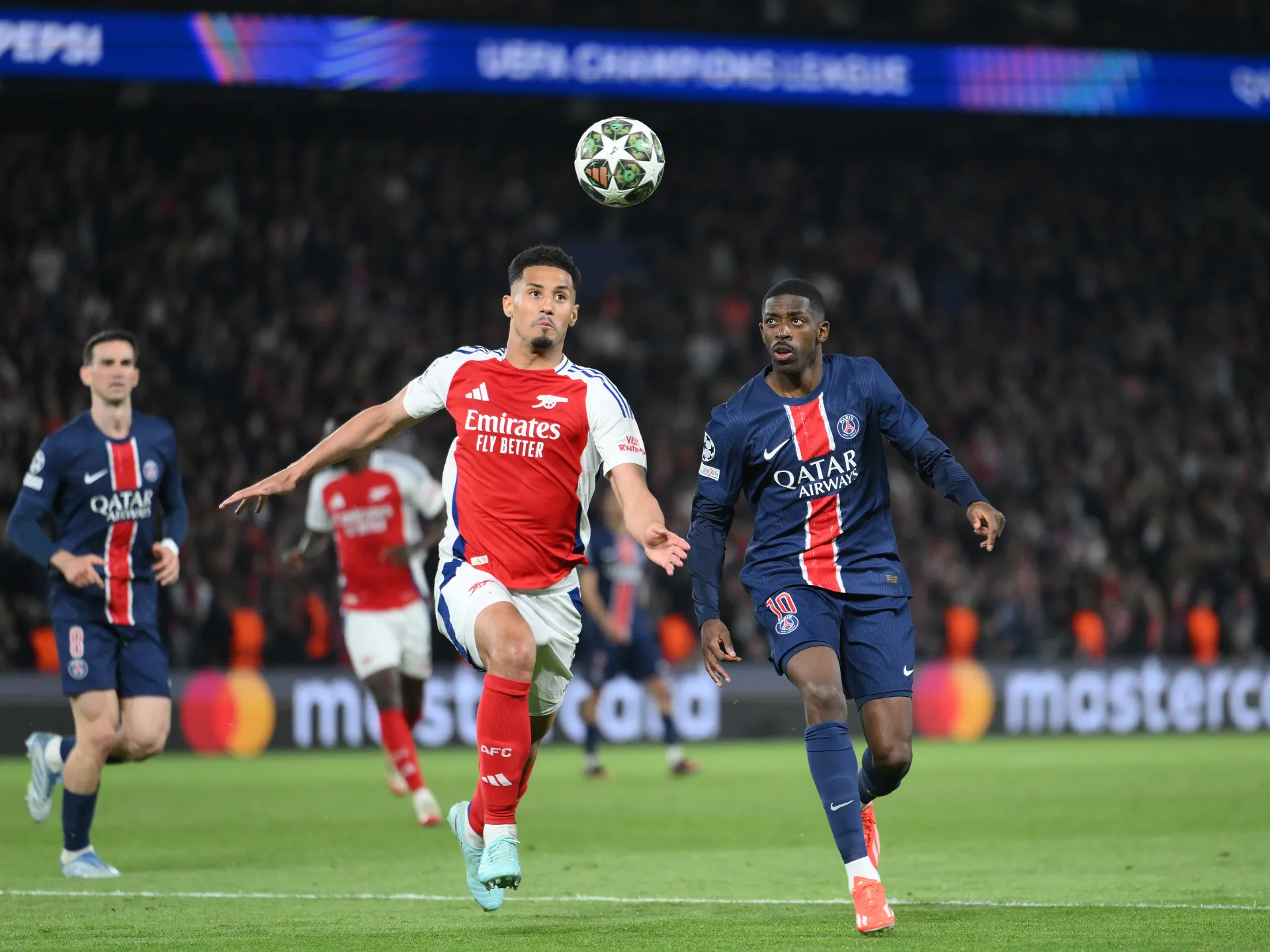 Soccer players chase a ball midair during a match between Arsenal and PSG with a crowded stadium in the background