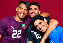 Three male soccer players hug and smile, wearing Moroccan team jerseys (purple, dark, and blue) with numbers visible, against a red background.