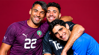 hjg | MCG24 | موقع إلكتروني مغربي شامل متجدد على مدار الساعة Three male soccer players hug and smile, wearing Moroccan team jerseys (purple, dark, and blue) with numbers visible, against a red background.