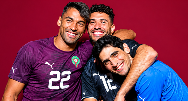Three male soccer players hug and smile, wearing Moroccan team jerseys (purple, dark, and blue) with numbers visible, against a red background.