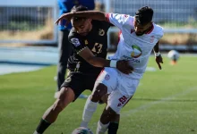Two soccer players contest the ball on a sunny green field, one in a black kit and one in white with a sponsor logo.