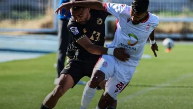 Two soccer players contest the ball on a sunny green field, one in a black kit and one in white with a sponsor logo.