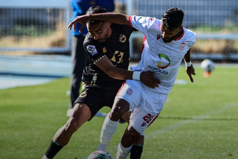 Two soccer players contest the ball on a sunny green field, one in a black kit and one in white with a sponsor logo.